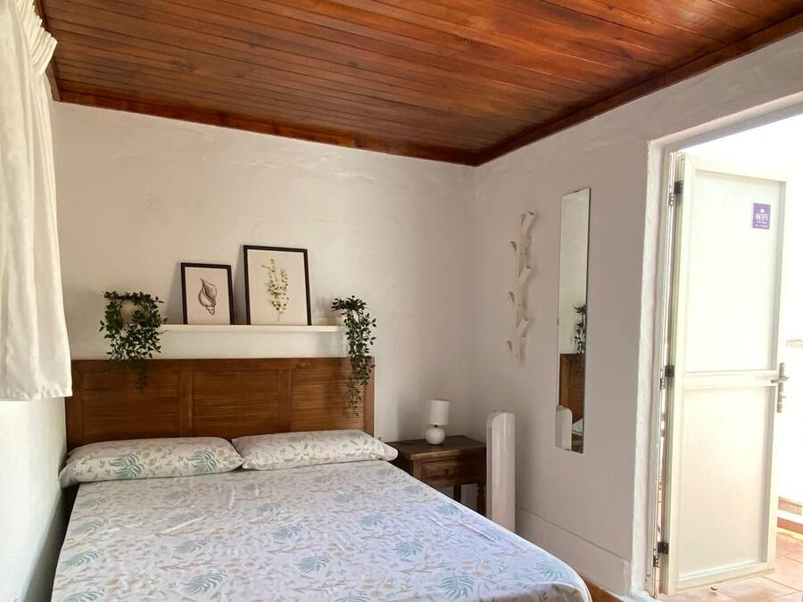 Une chambre avec un plafond en bois, un lit recouvert d’une couvre-lit à motifs floraux, une table de chevet, un miroir fixé au mur et une porte ornée du symbole du Wi-Fi.