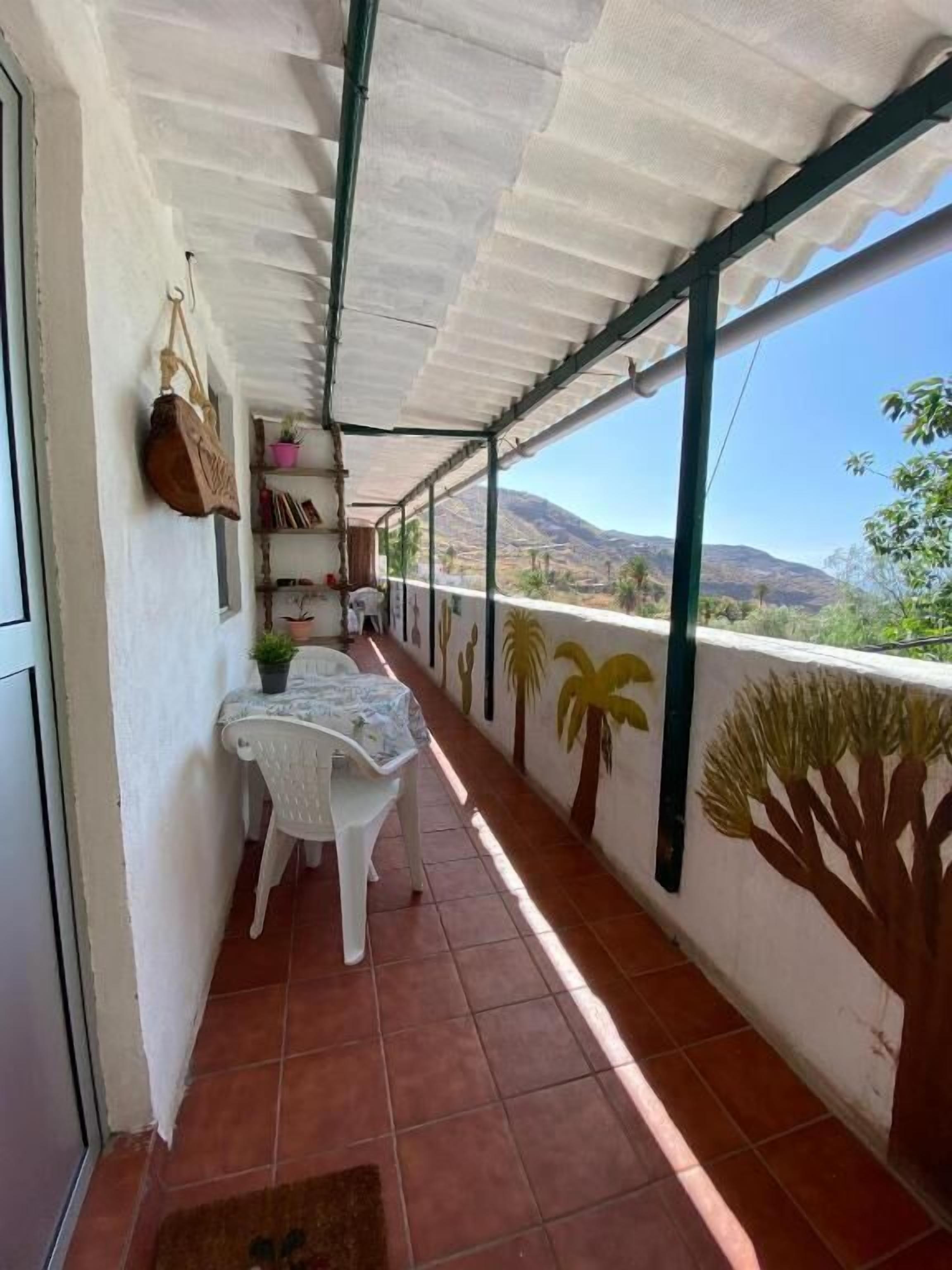 A covered balcony with a tiled floor, a white table with chairs, a hanging bag, and a shelf with books and a plant.