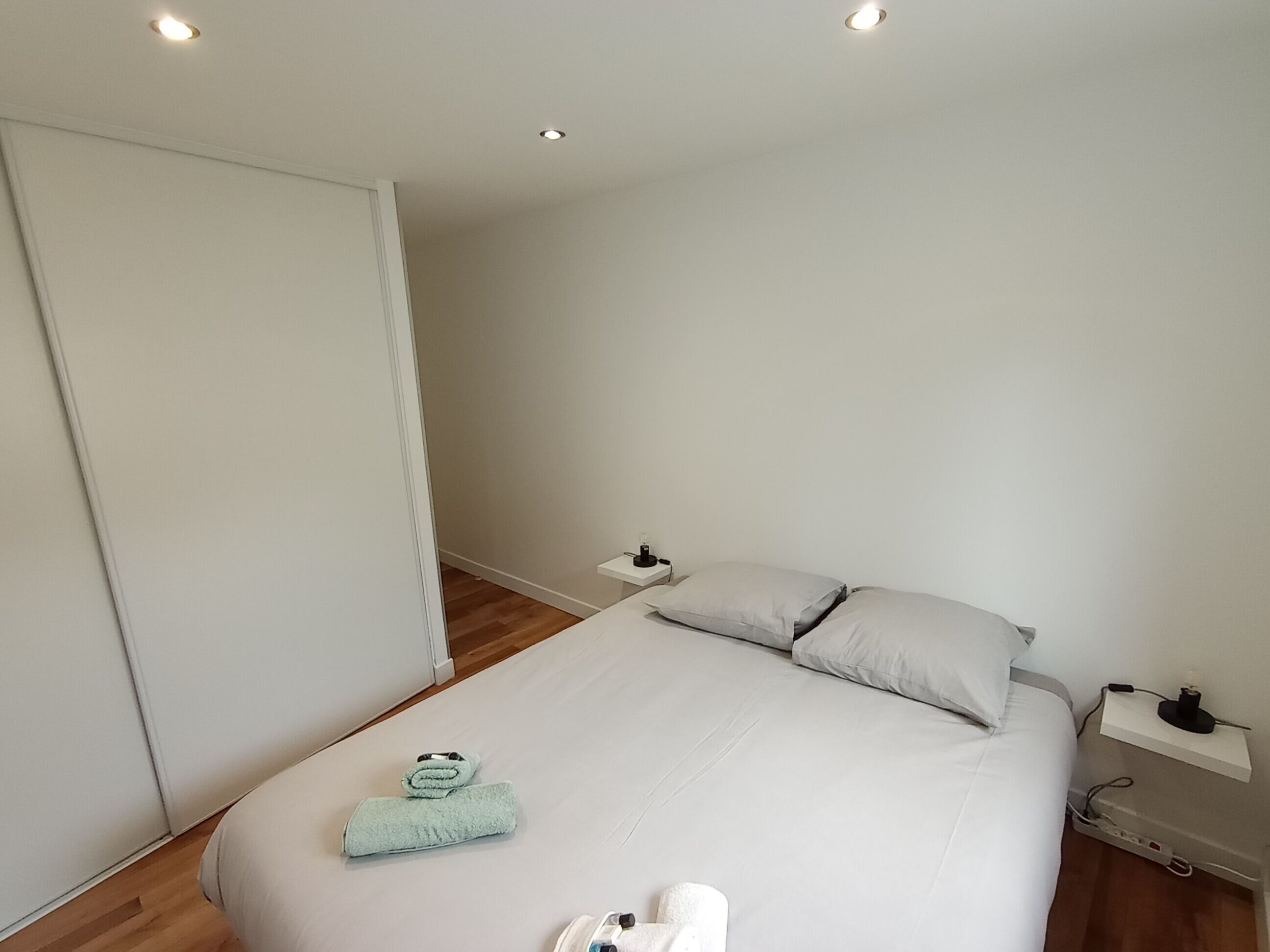 A bedroom with a bed, a nightstand, a bedside table, a towel, and a wardrobe.