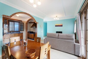 A dining area with a wooden table, chairs, and a basket. There is a kitchen with a brick archway and a sofa in the living area.