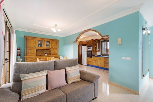 A living room with a sofa, a kitchen in the background, and a tiled floor.