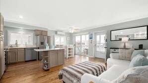 A modern kitchen with a central island, a dining area with a white table, and a living room with a sofa and a TV. - Nicely Renovated Two Bedroom Oceanside Condo (Ocean City)