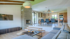 Un salon moderne avec un canapé, une table basse et un téléviseur fixé au mur. Un escalier mène à un espace repas avec des chaises, et on aperçoit une cuisine au fond.