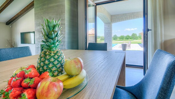 Un coin repas avec une table en bois, des fruits et une vue sur une piscine et de la verdure.