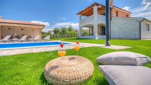 Un espace piscine avec des chaises longues, un pouf en osier et un bâtiment en pierre doté d’un balcon.