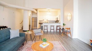 A modern living room with a sofa, coffee table, and a dining area with chairs and a small table. A kitchen is visible in the background with a bar counter and stools. - 3BR CozySuites TheBlock Skybridge Access (Indianapolis)