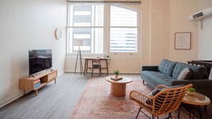 A living room with a TV, sofa, coffee table, and a large window. - 3BR CozySuites TheBlock Skybridge Access (Indianapolis)