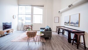 A spacious living room with a large window, a sofa, a coffee table, a TV, and a long table with a board game. - 3BR CozySuites TheBlock Skybridge Access (Indianapolis)