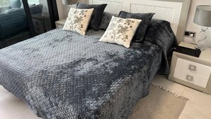 A bed with a textured grey quilt, two pillows with floral patterns, a bedside table with a lamp, and a view through a glass door. - Apartment 'Sunset Waves' with Sea View, Wi-Fi and Air Conditioning (Benidorm)