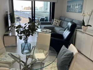 Una sala de estar moderna con una mesa de centro de vidrio, un sofá gris y vistas de la ciudad a través de amplias ventanas.
