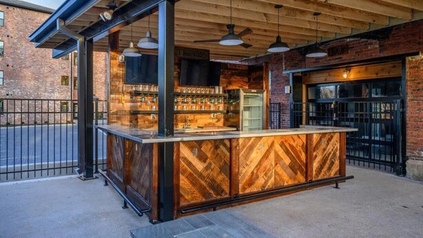Un área de bar al aire libre con mostrador de madera, luces colgantes y pared de ladrillo de fondo.