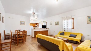 A room with a sofa, a dining table with chairs, a kitchen area, and a TV.