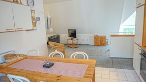 Un coin repas avec une table en bois, des chaises blanches, une nappe à carreaux rouges, un téléviseur posé sur un meuble, une chaise en osier et un buffet avec une porte en bois.