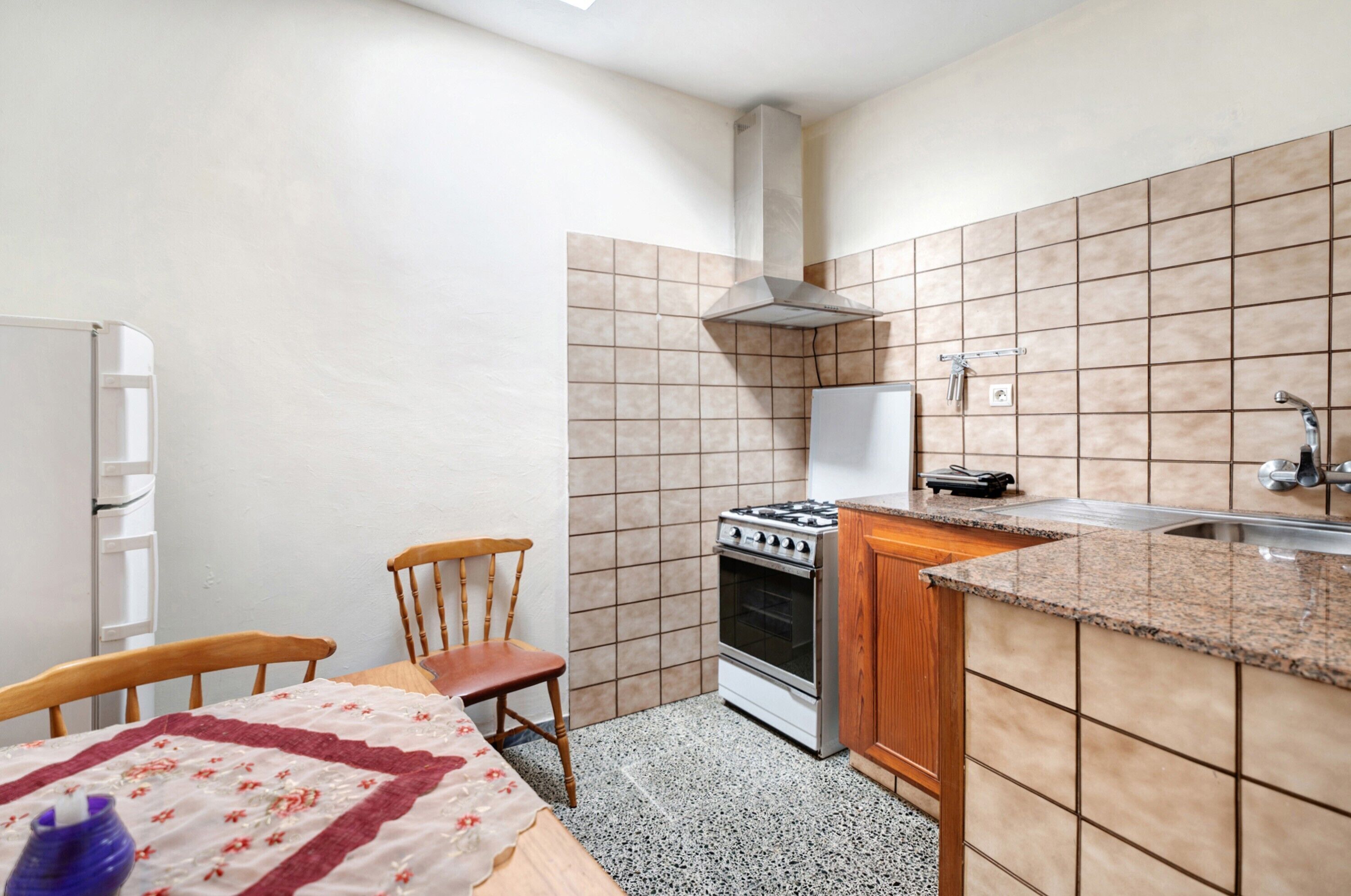 A compact kitchen with a stove, oven, and sink, a wooden chair, and a table with a tablecloth.