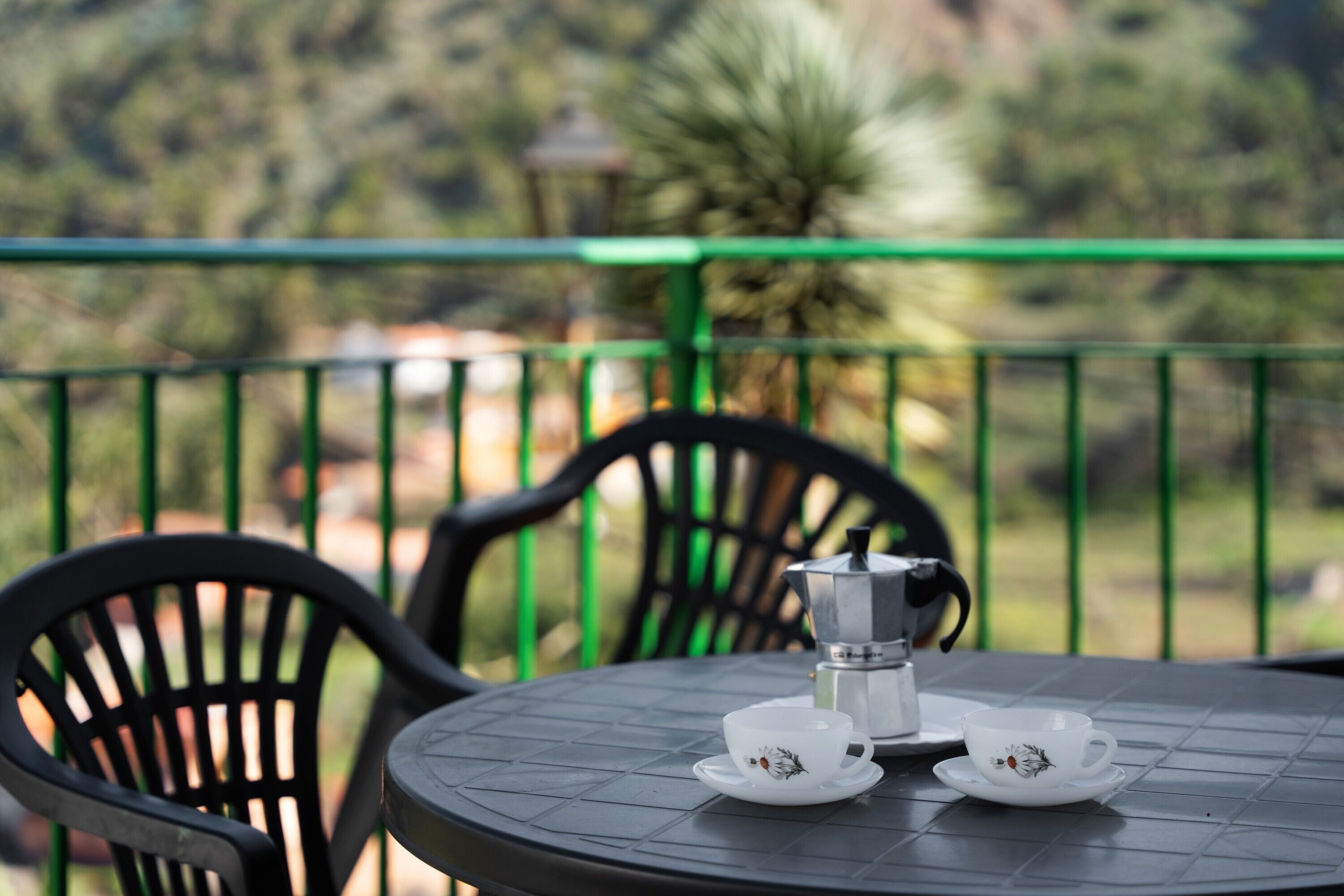 A round table with two white cups and a coffee pot, surrounded by black chairs, on a balcony with a green railing.