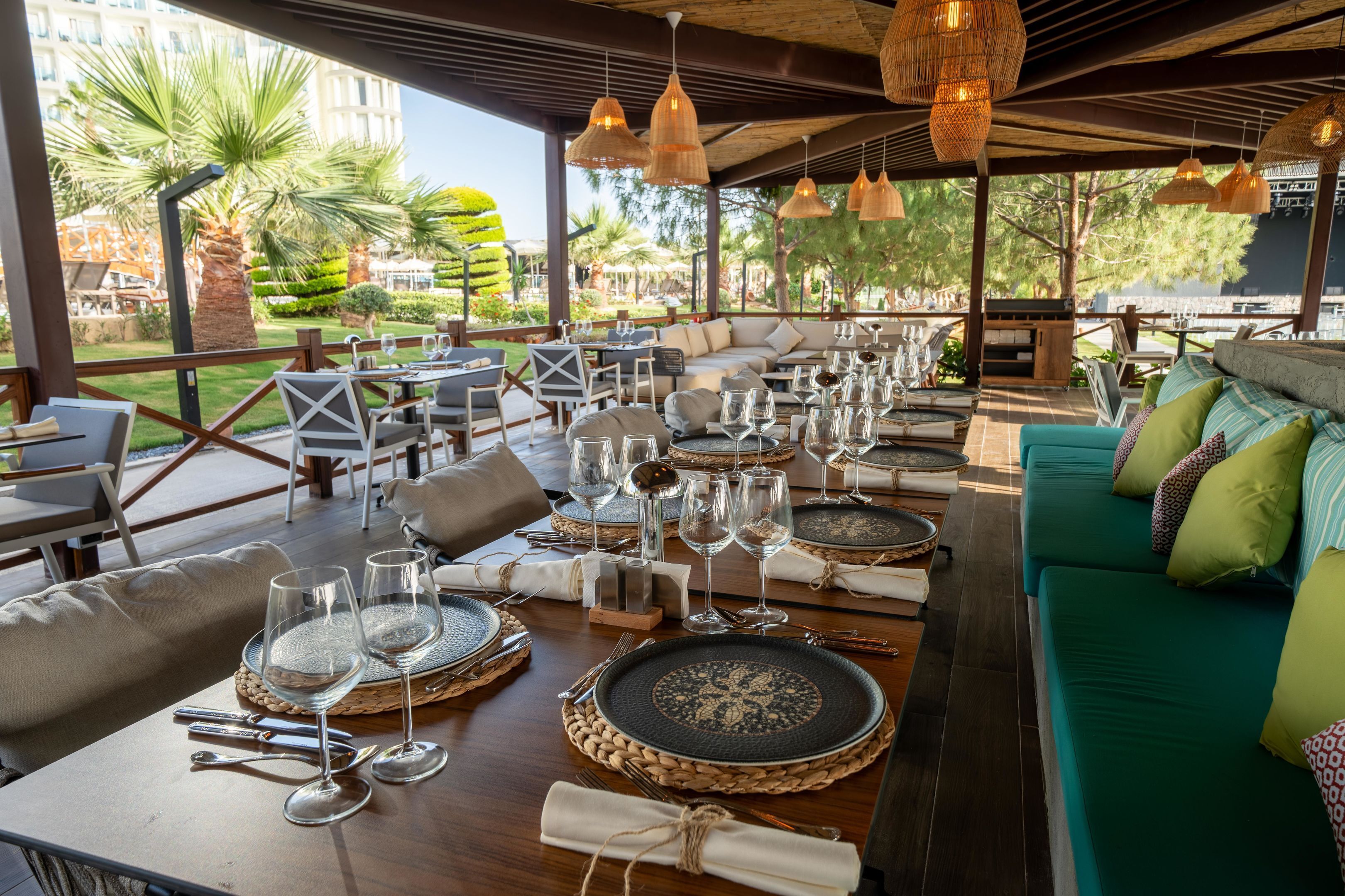 An outdoor dining area with tables set for a meal, surrounded by greenery and a view of a building.