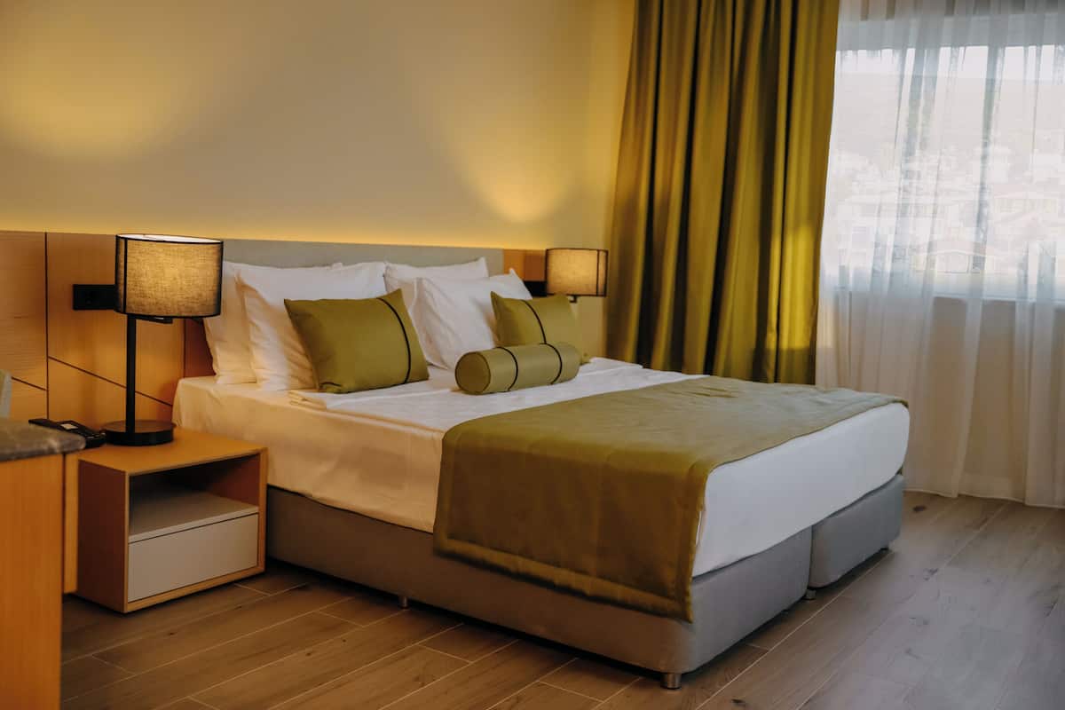 Une chambre d’hôtel moderne avec un grand lit, deux tables de chevet, une petite table et une fenêtre avec des rideaux.