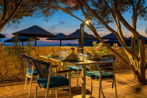 Un área de comedor al aire libre con un muro de piedra, terraza de madera y sombrillas de paja.