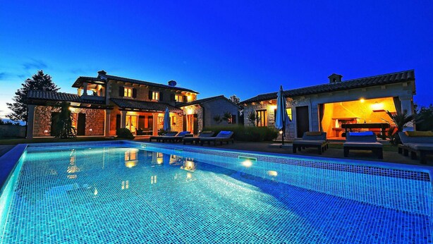 Une grande piscine avec une terrasse carrelée, entourée d’un bâtiment en pierre comprenant plusieurs pièces et un espace bar.