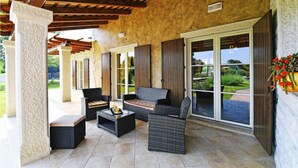 Un espace extérieur aménagé en terrasse, avec des meubles en osier, un sol en pierre et une vue sur un jardin, visible à travers de grandes portes-fenêtres coulissantes.