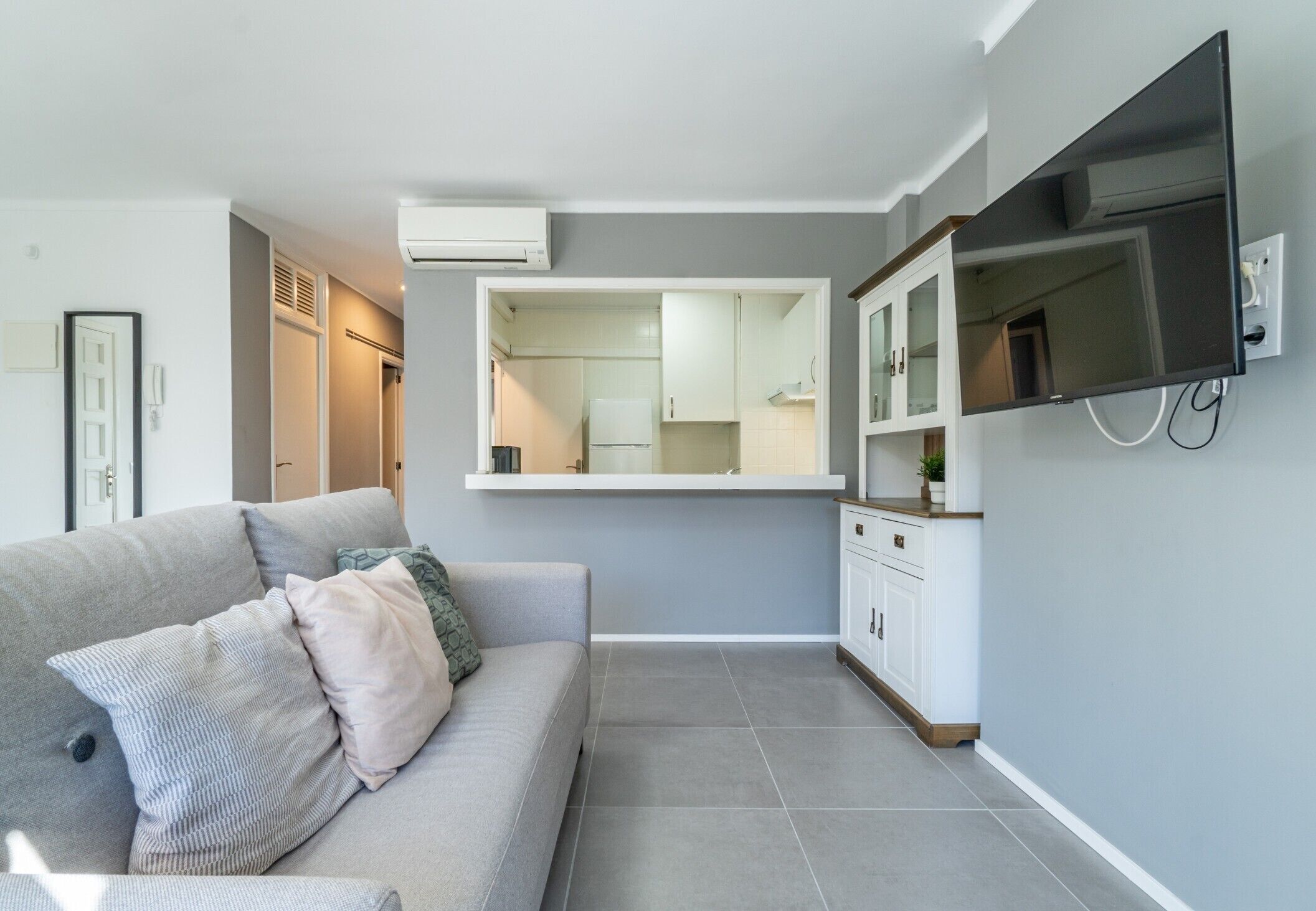 Una sala de estar moderna con un sofá, un televisor de pantalla plana montado en la pared y una cocina visible a través de un puerta abierta.