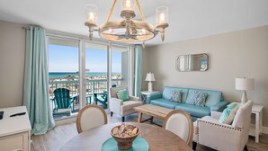 Een ruime woonkamer met een groot raam met uitzicht op het strand, een kroonluchter, een blauwe bank en een eettafel met een schaal fruit.