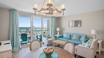 A spacious living room with a large window overlooking the beach, a chandelier, a blue sofa, and a dining table with a bowl of fruit.