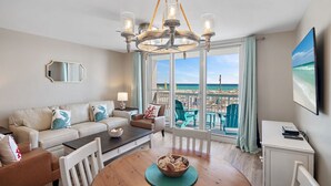 A modern living room with a view of the beach, a chandelier, a dining table, and a television. - Renovated beachfront condo, spectacular Gulf views (Destin)