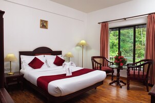 Un cuarto de hotel con una cama grande, dos mesitas de noche con lámparas, una mesita pequeña con un florero y vista a la vegetación por la ventana.