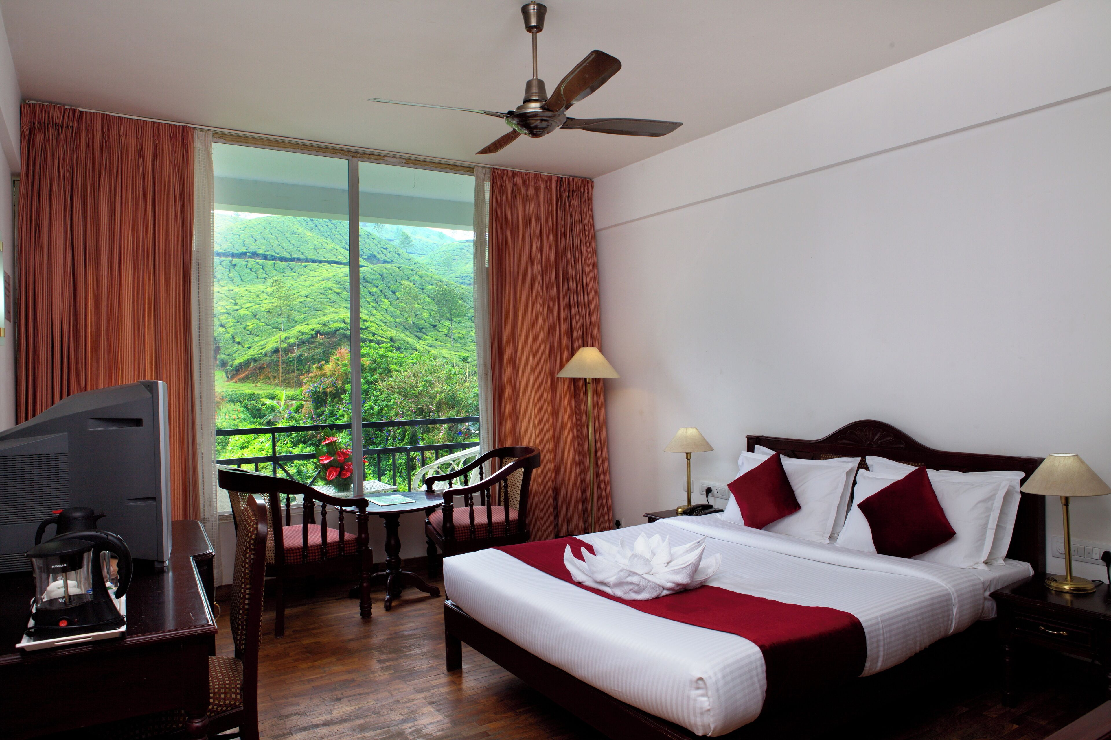 Habitación de hotel con cama, escritorio con televisor, una silla, una mesa y vista a un paisaje verde exuberante a través de amplios ventanales.