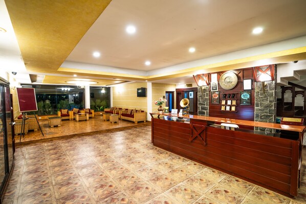 A reception area with a wooden reception desk, a seating area with sofas, and a tiled floor. - Hotel C7 Munnar (Devikolam)