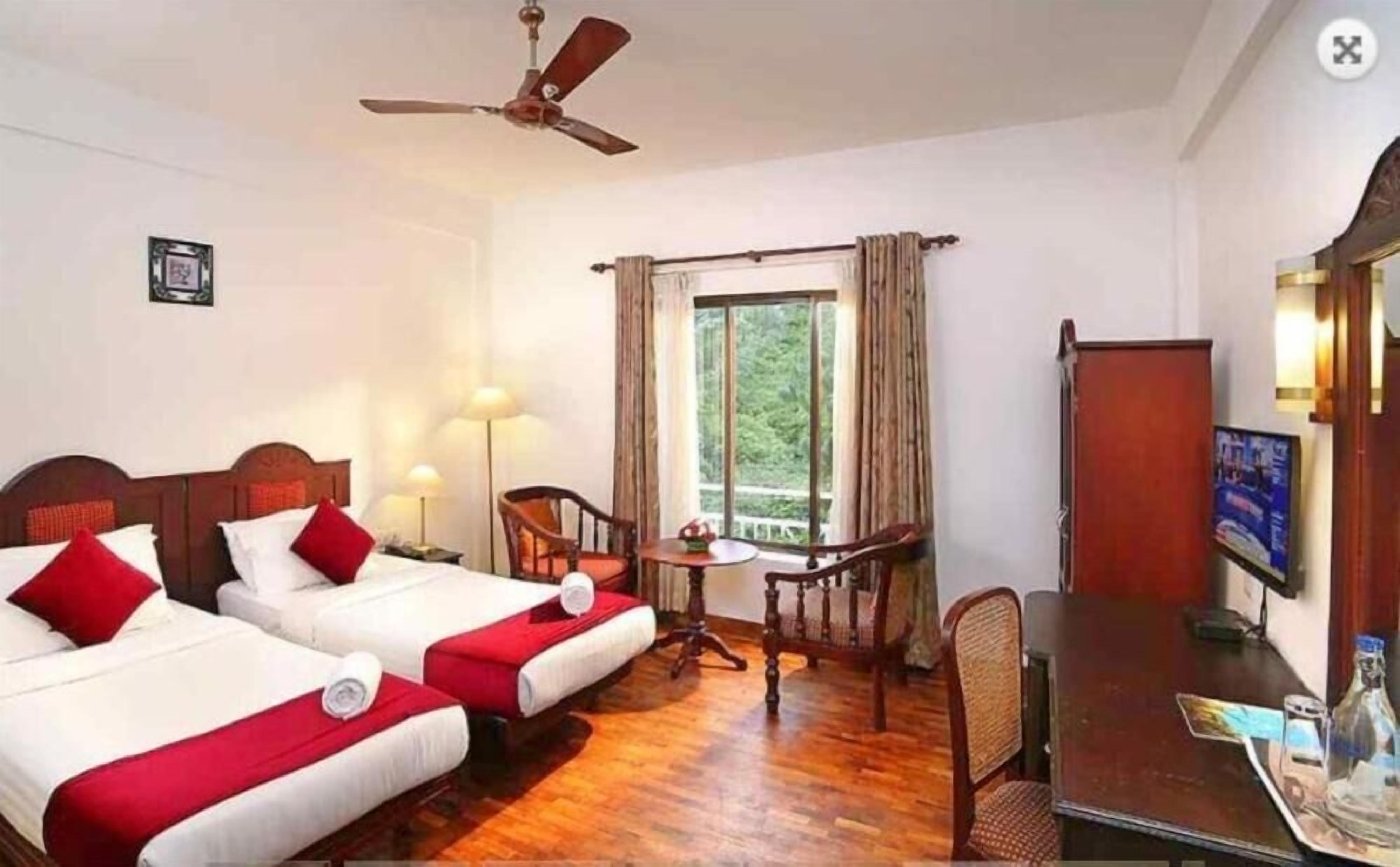 A hotel room with two beds, a ceiling fan, a small table with chairs, a TV, and a window with curtains.