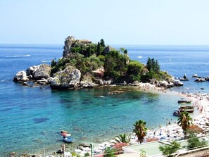 Une vue sur la côte, avec une île rocheuse, une eau d’un bleu limpide et une plage parsemée de parasols et de bateaux.