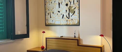 A bedroom with a bed, a wall-mounted artwork, bedside tables with lamps, and a window with shutters.