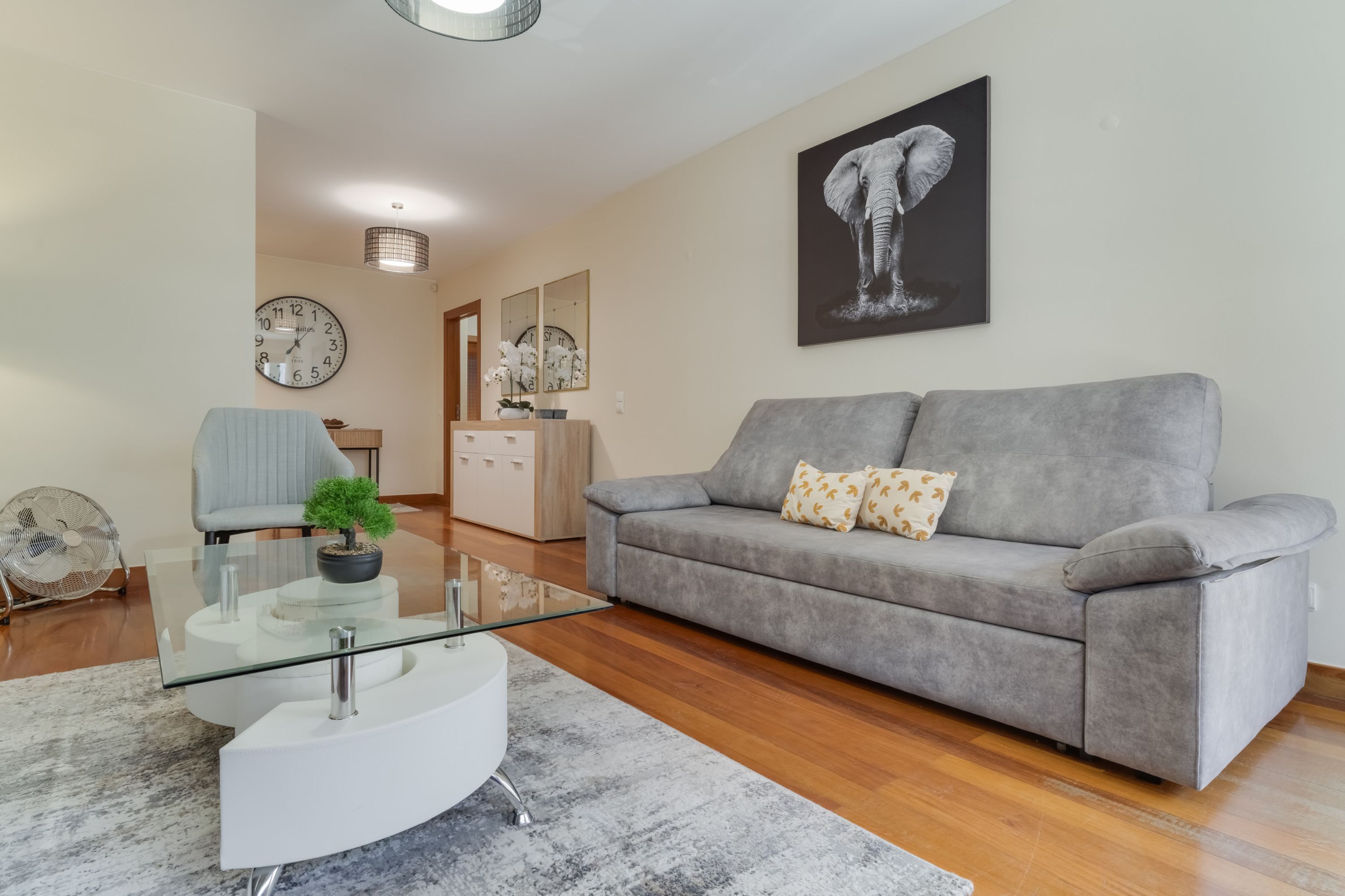 A modern living room with a grey sofa, a glass coffee table, a white side table, a fan, and a clock on the wall.