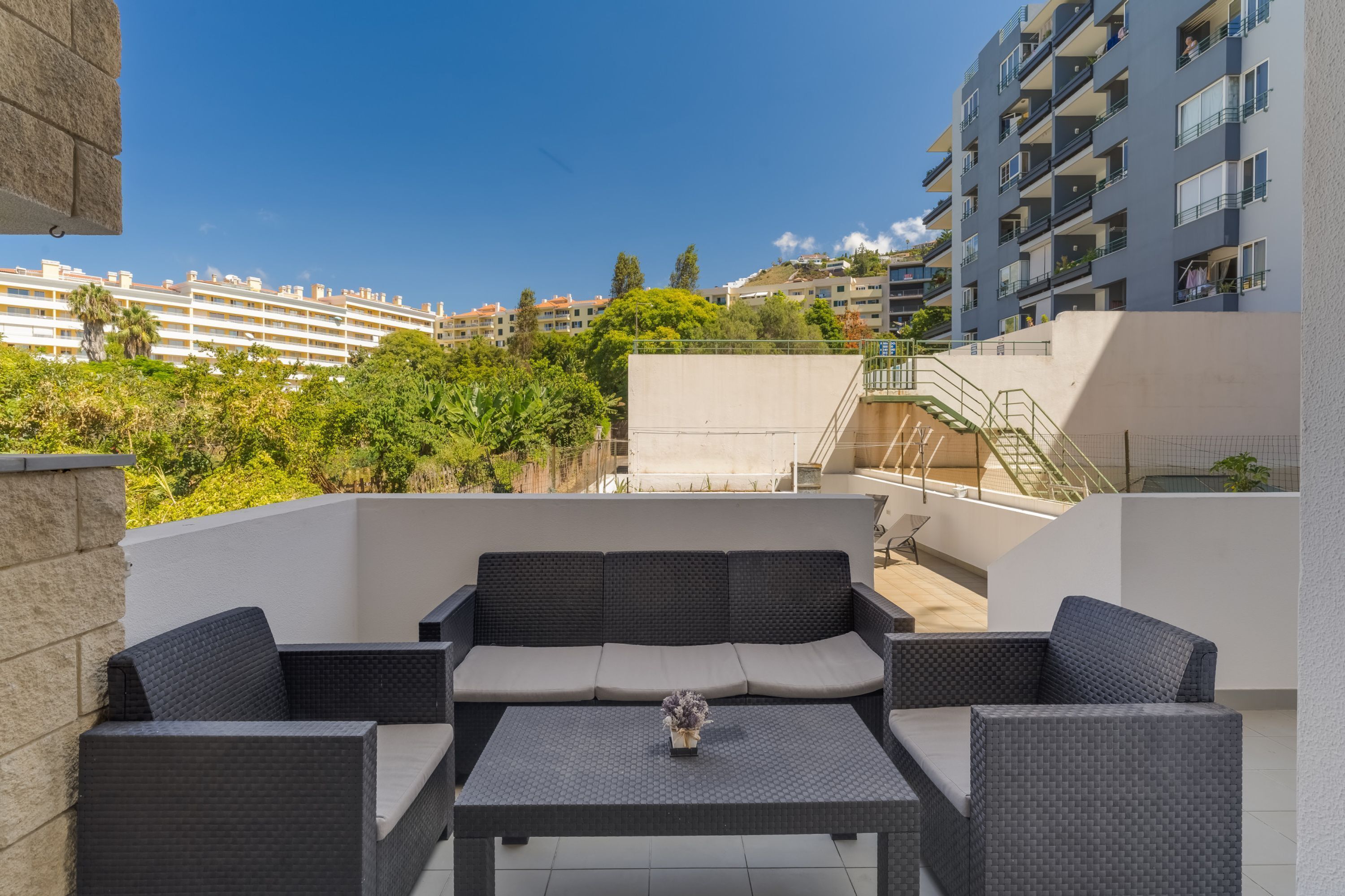 Una terraza en la azotea con mobiliario exterior, vistas de edificios circundantes y una escalera que lleva a un nivel superior.
