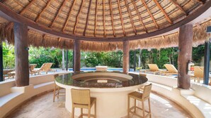 Un espace bar circulaire doté d’un toit de chaume, entouré de fauteuils et d’une piscine.