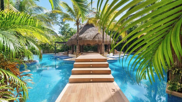 Une piscine avec une terrasse en bois menant à une structure à toit de chaume, le tout entouré de palmiers.
