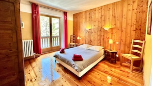 Une chambre aux murs lambrissés, comprenant un lit, deux lampes de chevet, un fauteuil et une vue sur l’extérieur, accessible par une porte-fenêtre coulissante.