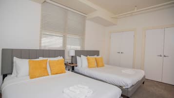 Habitación de hotel con dos camas, ropa de cama blanca y almohadas amarillas. Hay dos puertas y una ventana con persianas.