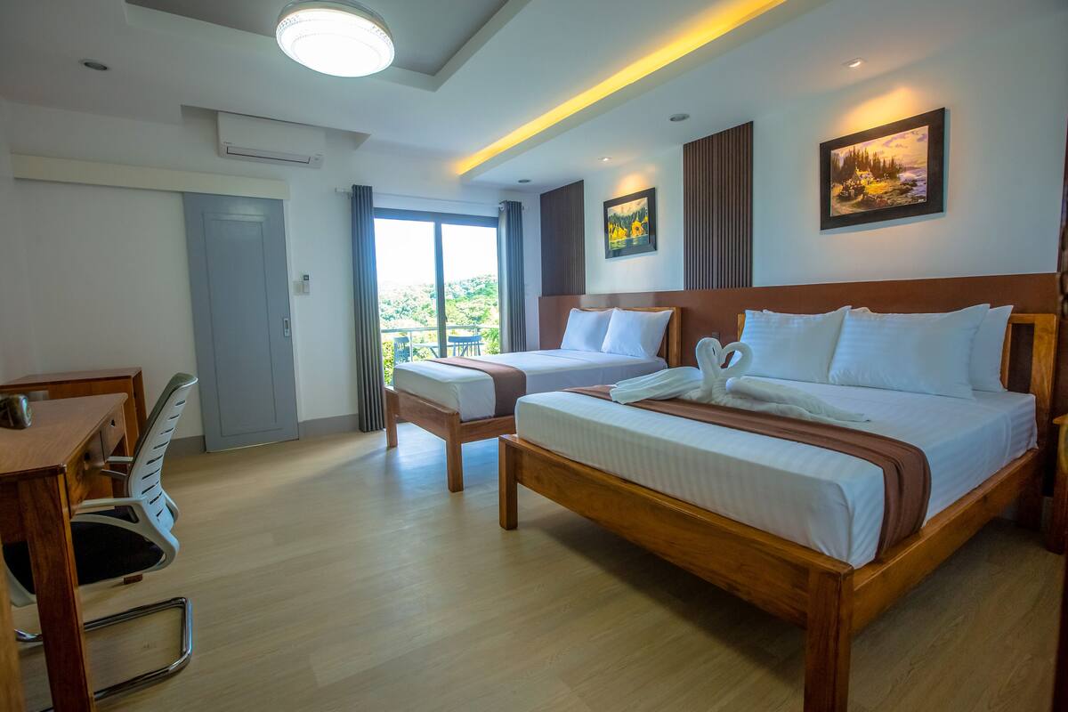 A hotel room with a large bed, a desk, and a chair. There is a balcony with a view of the outdoors.
