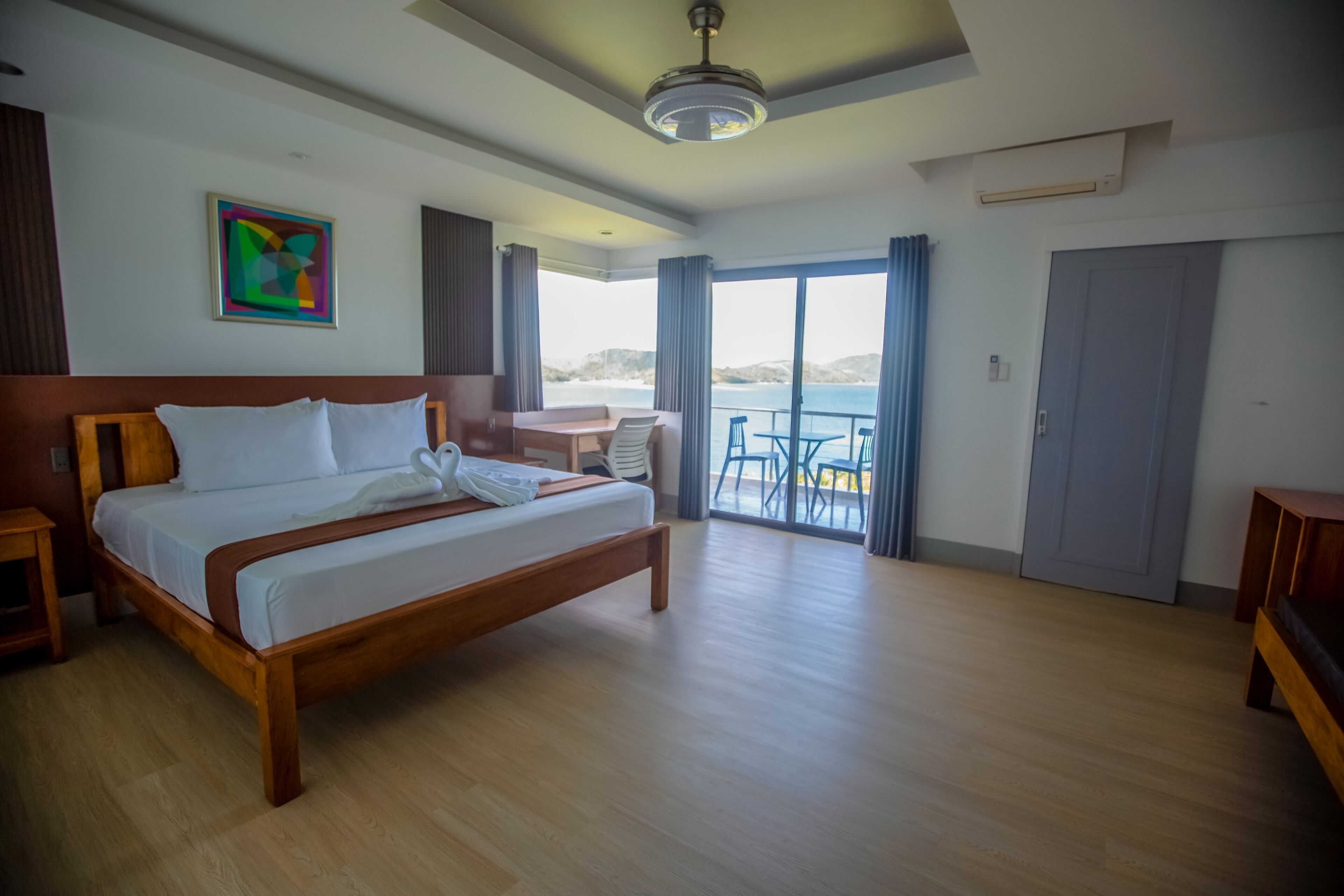 Une chambre d’hôtel avec un lit en bois, une petite table, une chaise et une vue sur la mer grâce à de grandes fenêtres.