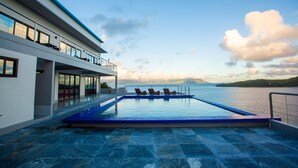 Un área de piscina moderna con vistas al mar, una terraza techada con mobiliario y un cielo despejado.