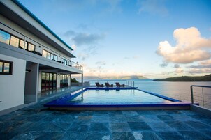 Un área de piscina moderna con vistas al mar, una terraza techada con mobiliario y un cielo despejado.