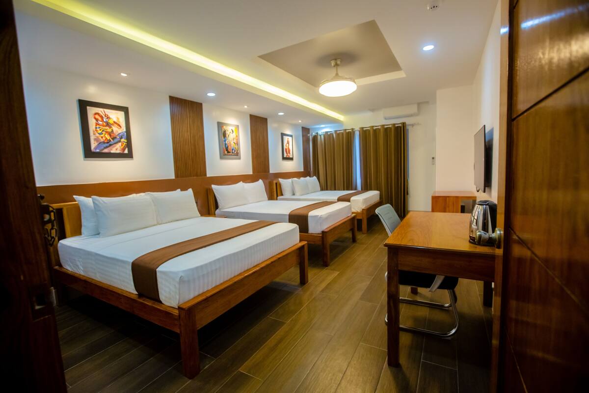 A hotel room with three single beds, a wooden desk, a television, and artwork on the walls.
