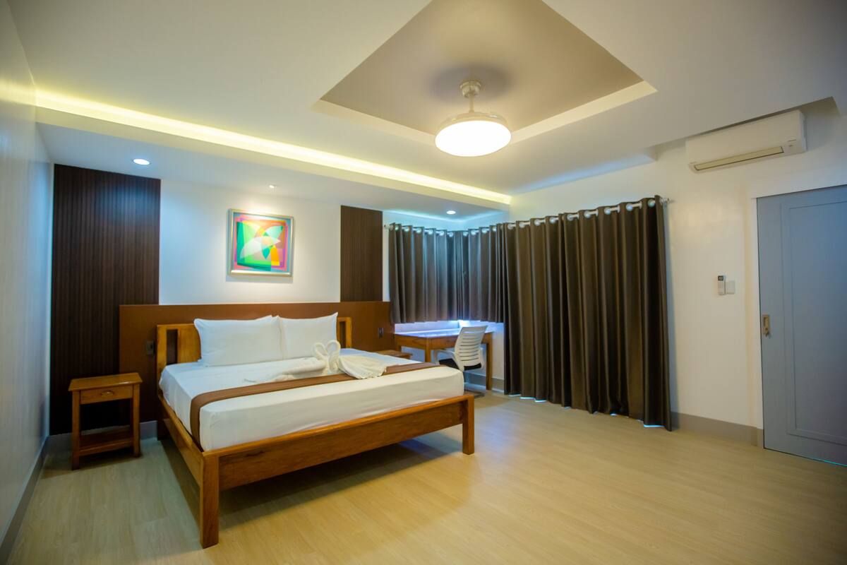 A modern hotel room with a wooden bed, bedside tables, a desk, and a large window with curtains.