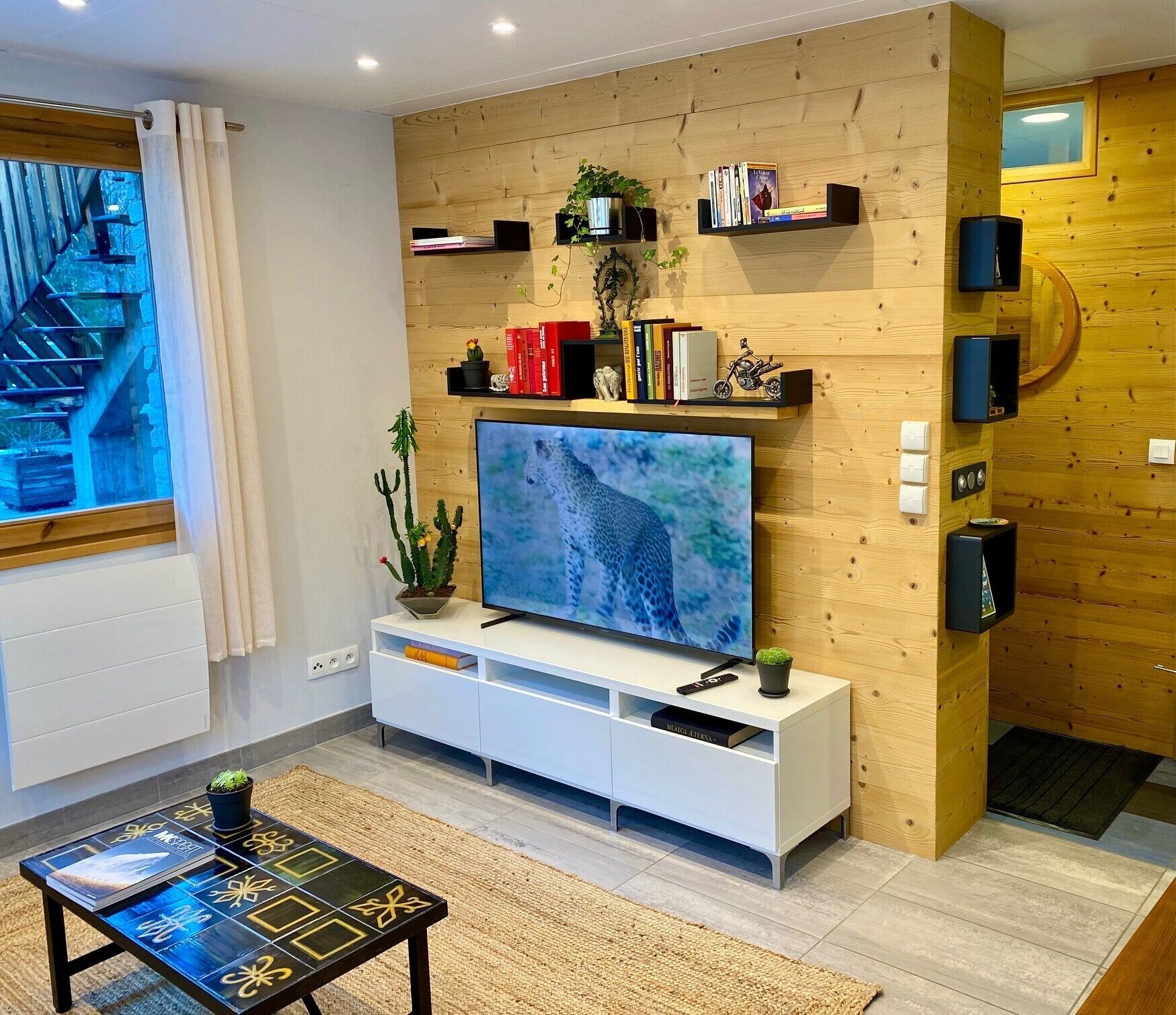 Ein modernes Wohnzimmer mit Fernseher, Holzregalen mit Büchern und Dekoration, einem kleinen Couchtisch und Blick auf eine Treppe.
