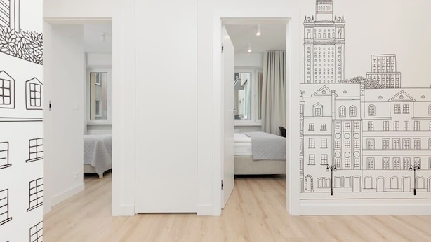 Un pasillo con una pared blanca y un mural que representa un paisaje urbano. Se ven dos dormitorios, cada uno con una cama y una ventana.
