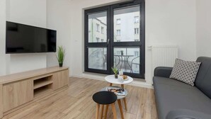 Un salon moderne doté d’un téléviseur à écran plat, d’un meuble de rangement en bois, d’une table basse ronde agrémentée d’une plante, d’un tabouret noir et d’un canapé gris orné de coussins à motifs.