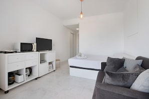 Une chambre moderne avec un bureau blanc, un téléviseur à écran plat, un lit avec un matelas et des oreillers, et un canapé gris.